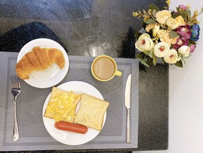 A breakfast setting with croissant, scrambled eggs, toast, and a hot dog on a plate, a cup of coffee, and a bouquet of flowers. - Okaeri Homestay (Hanoi)