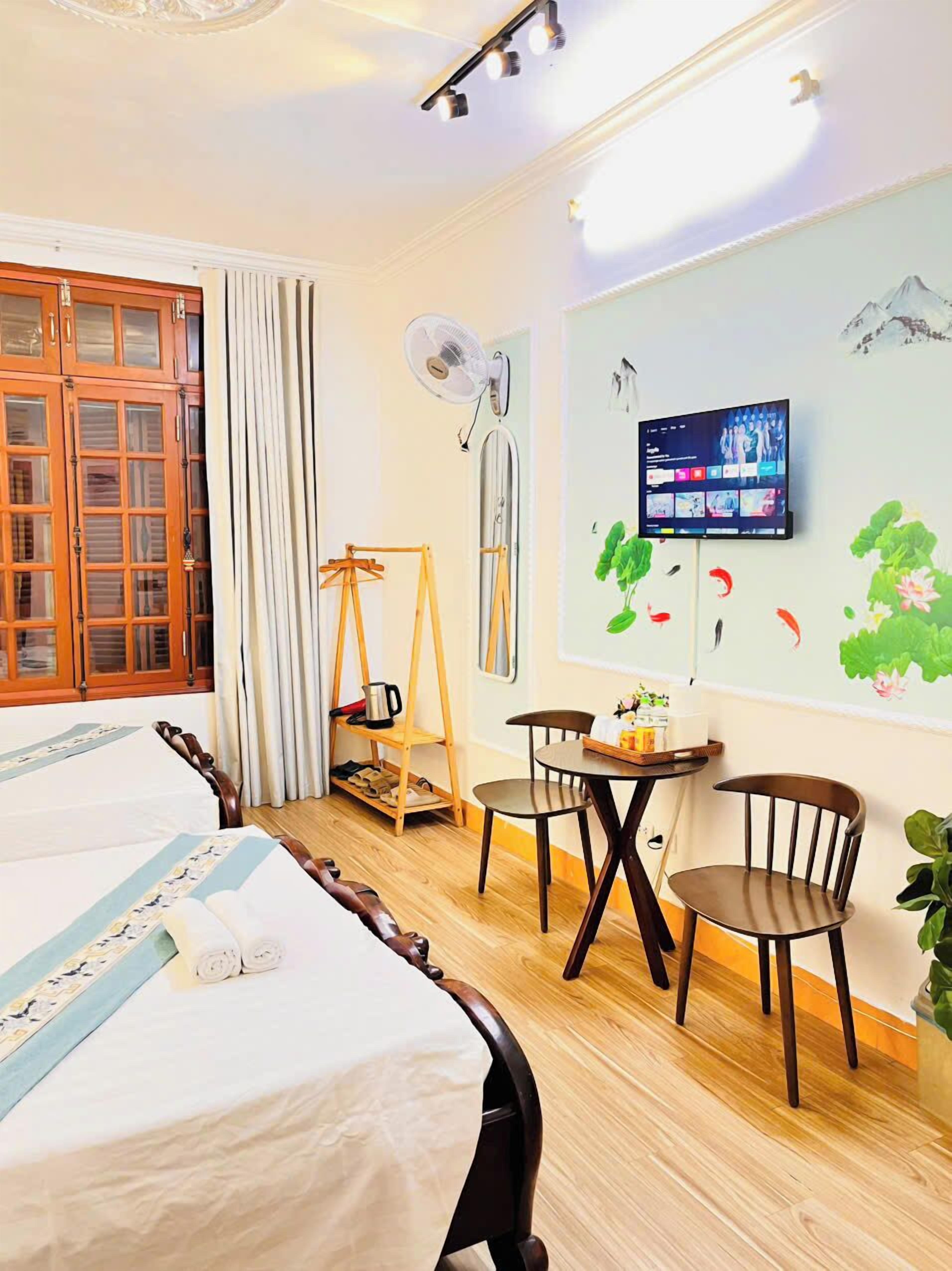 A hotel room with a bed, two chairs, a small table, a fan, a TV, and a window with curtains.