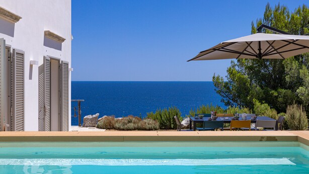 Una piscina con vista al mar, mobiliario exterior y un edificio con persianas.