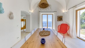 Un comedor con una mesa de madera, dos jarrones, una lámpara colgante de fibras naturales, una silla roja y vistas al mar a través de un ventanal grande.