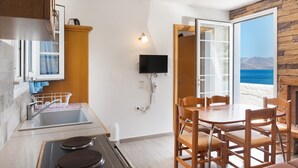 Eine Küche mit Spüle, Herd und Essbereich mit Stühlen. Ein Fernseher ist an der Wand befestigt und durch eine Tür bietet sich Blick auf das Meer.