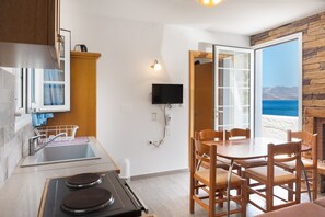 Une cuisine équipée d’un évier, d’une cuisinière et d’un coin repas avec des chaises. Un téléviseur est fixé au mur et, en passant par la porte, on peut admirer la vue sur la mer.