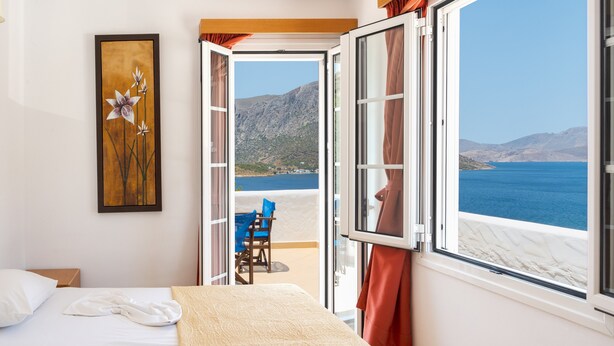 Ein Hotelzimmer mit Bett, einem Bild, einer offenen Tür, die auf einen Balkon mit Blick auf Meer und Berge führt.