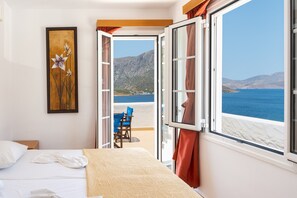 Une chambre d’hôtel avec un lit, un tableau, une porte ouverte donnant sur un balcon avec vue sur la mer et les montagnes.