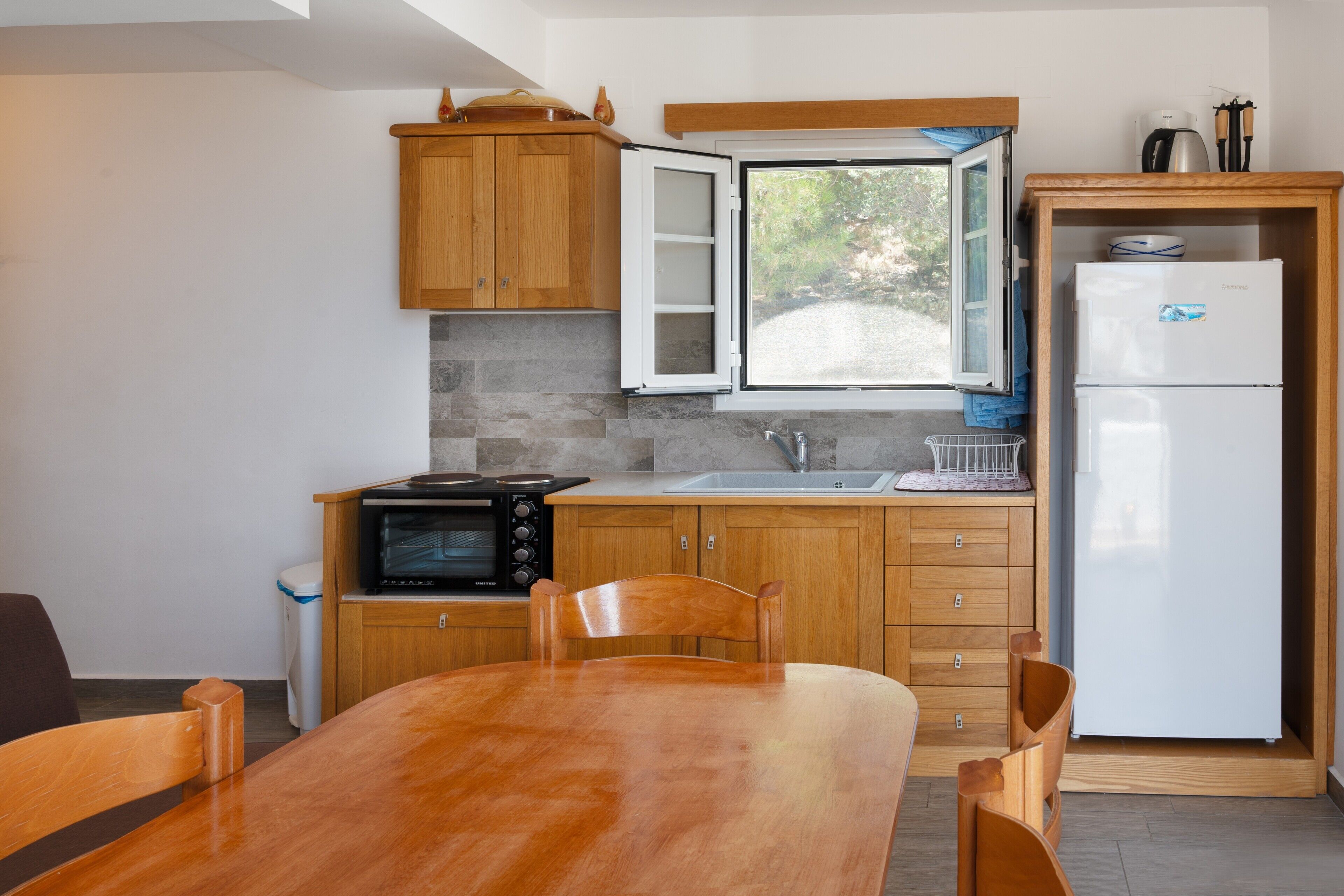 Una cocina con armarios de madera, microondas, refrigerador, mesa de comedor con sillas y ventana con vistas a zonas verdes.