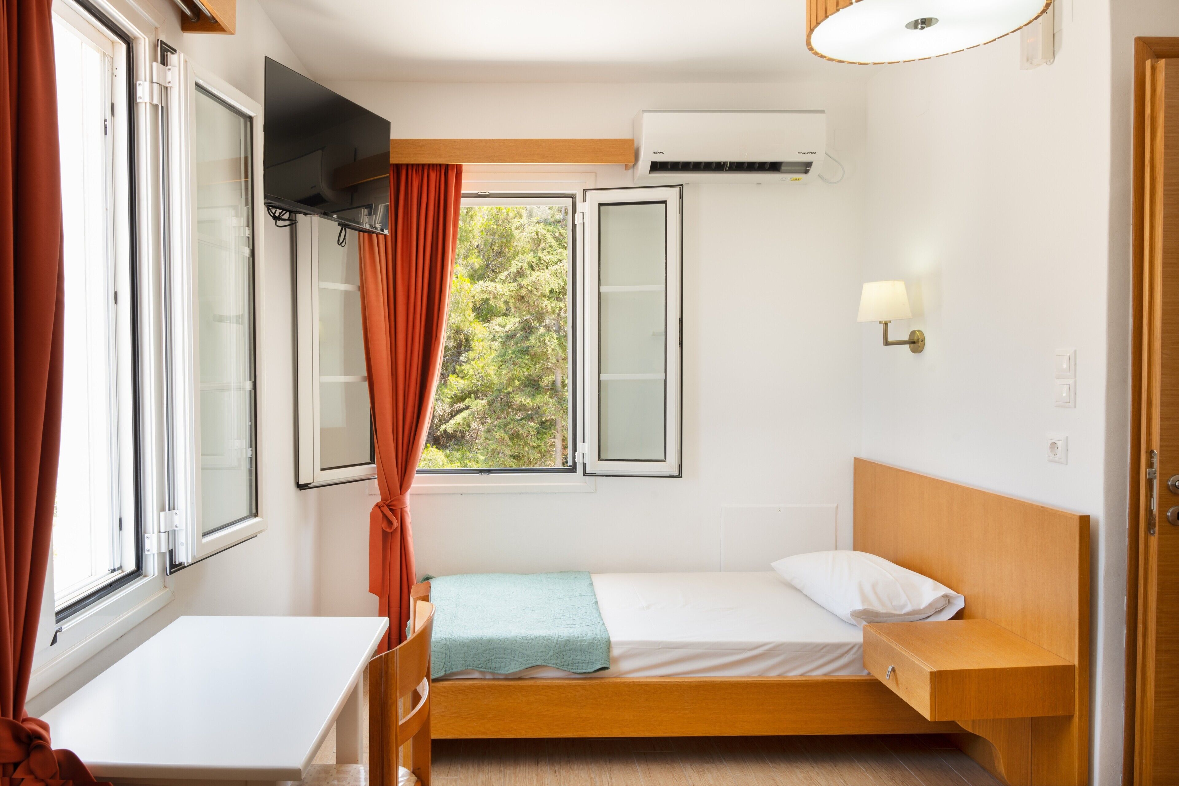 A single bed with a wooden headboard, a bedside table, a TV mounted on the wall, an air conditioning unit, and a window with curtains.