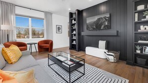 Een moderne woonkamer met een glazen salontafel, een bank met kussens, een witte fauteuil, een boekenkast en een schilderij aan de muur.