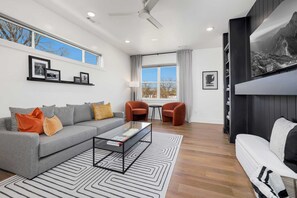 A modern living room with a grey sofa, orange and yellow cushions, a coffee table, and a patterned rug. There are two armchairs, a TV, and a bookshelf with framed pictures.