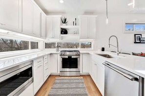 A modern kitchen with white cabinetry, a built-in oven, a dishwasher, and a window providing a view of trees.