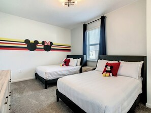 A hotel room with two beds, a Mickey Mouse wall decoration, and a window with curtains.