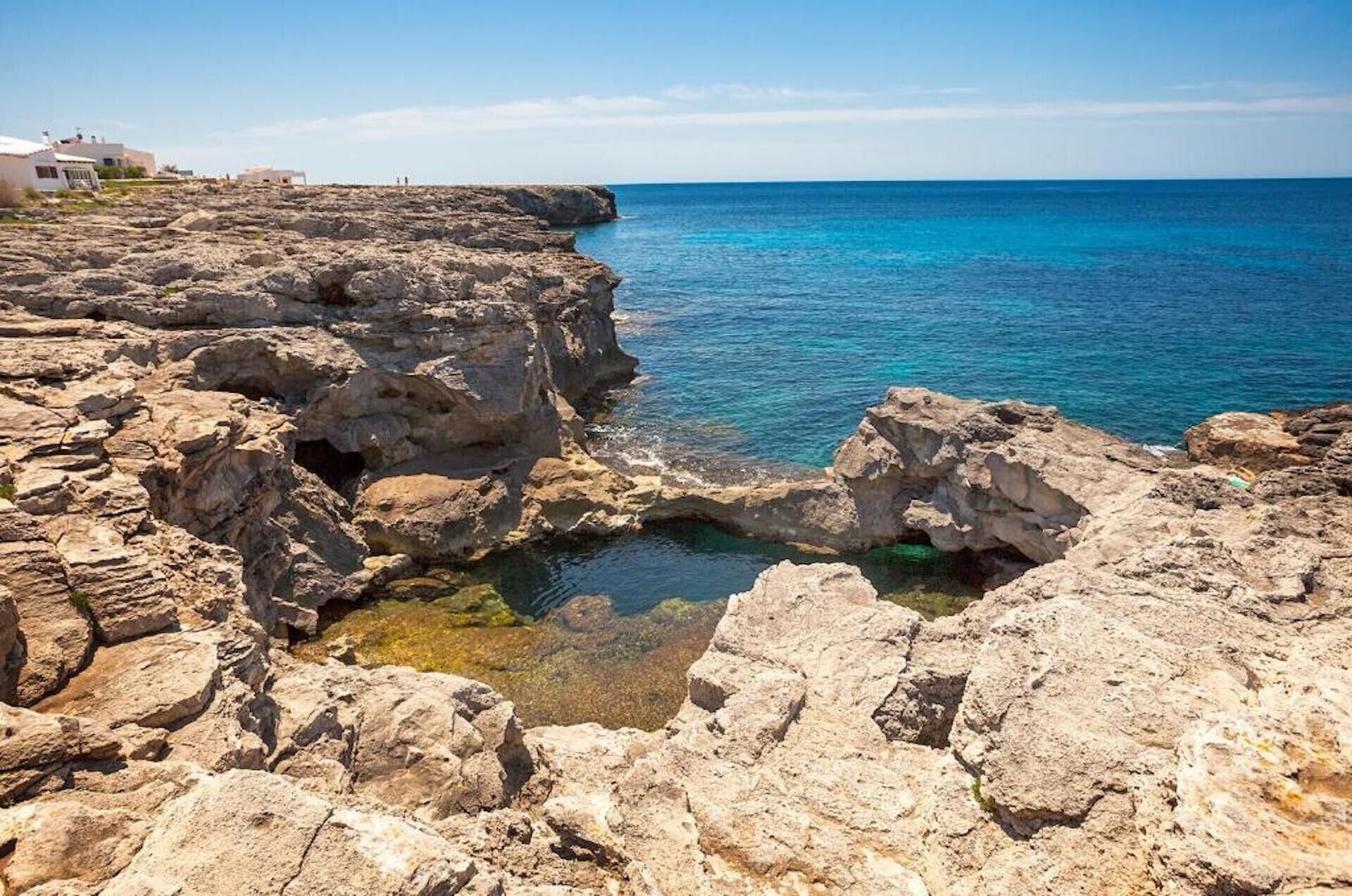 A rocky coastline with clear blue water, a small cave, and buildings in the background.