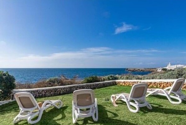 A seaside view with lounge chairs on a grassy area, overlooking the sea and a distant coastline. - Villa 'Bini Sek' with Sea View, Private Terrace and Air Conditioning (Binibèquer)