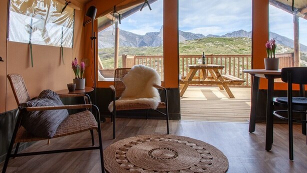 A cozy room with wicker chairs, a round rug, and a view of mountains through large windows.