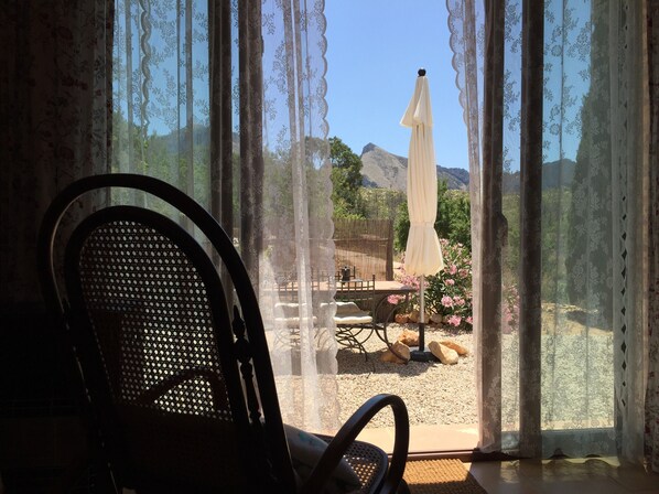 A room with a view of a garden through sheer curtains, a chair, and a table with an umbrella.