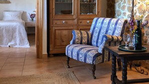 Une pièce comprenant un lit, un fauteuil à motifs, une armoire en bois avec des portes en verre, une table avec une lampe et un mur en pierre.