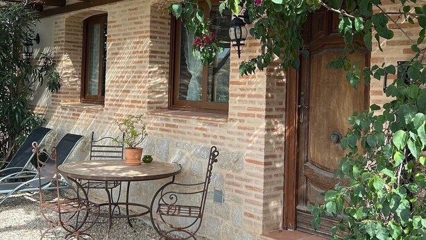 Un bâtiment rustique en pierre, doté d’une porte en bois, d’une fenêtre et d’une table avec des chaises à l’extérieur.