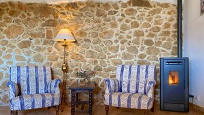 Un mur de pierres, deux fauteuils à motifs, une petite table avec une lampe et un poêle à bois.