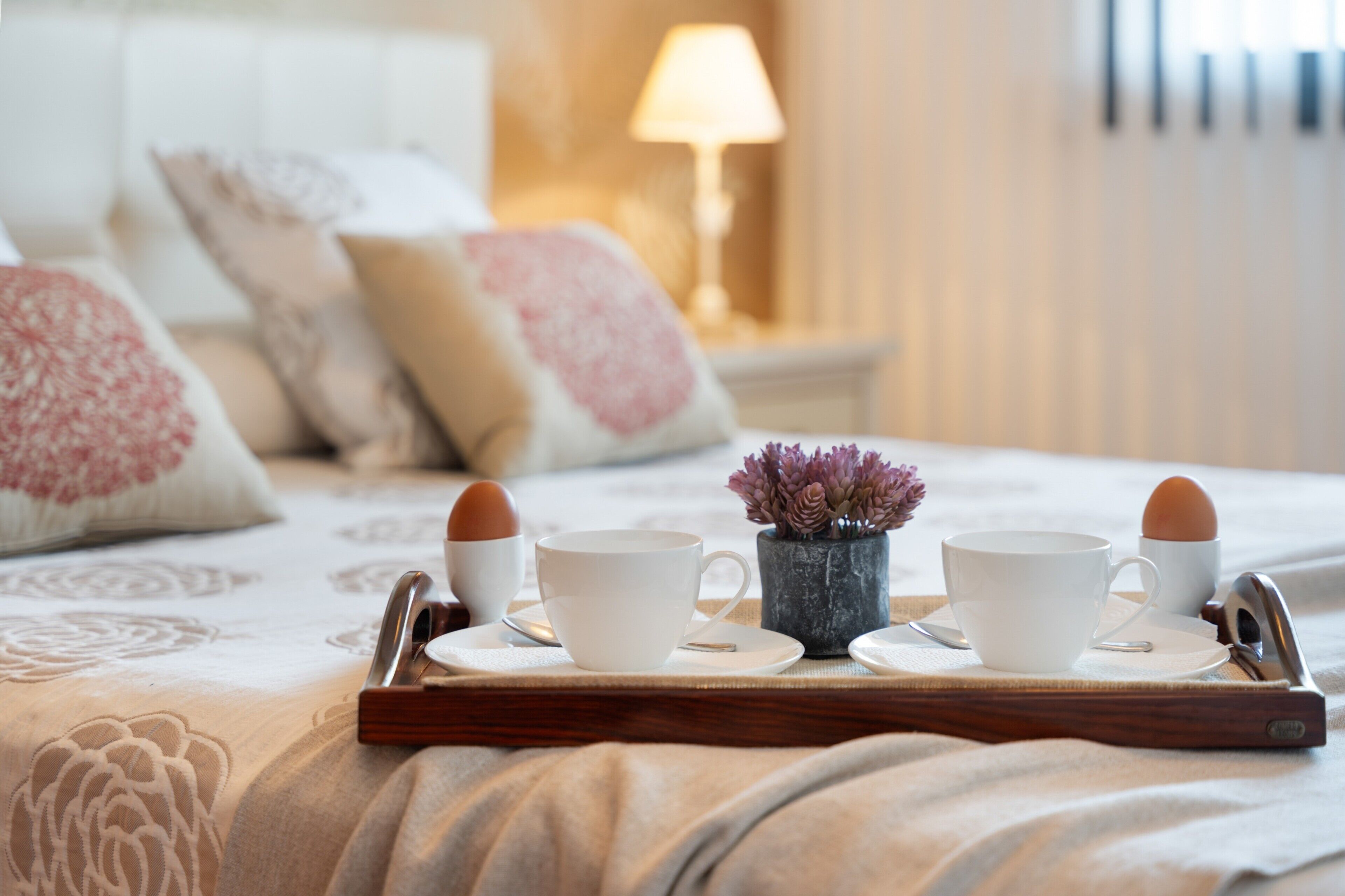 A breakfast tray with two cups and two eggs on a bed with a floral bedspread and pillows.