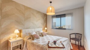 A bedroom with a bed, bedside tables, a chair, a window with curtains, and a city view. - Vacation Home "Vista Paraiso" with Sea View, Shared Pool & Wi-Fi (Adeje)