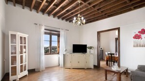 Un vaste salon doté d’un plancher en bois, d’un téléviseur posé sur un meuble, d’un lustre et d’un coin repas visible à travers une porte ouverte.