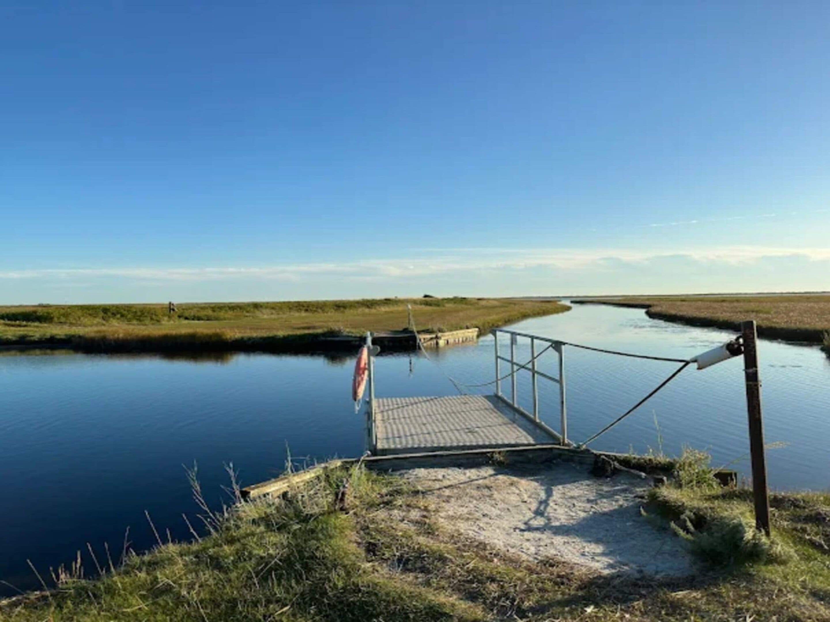 Un plan d’eau calme, avec une jetée en bois qui s’avance sur l’eau, entouré de zones herbeuses.