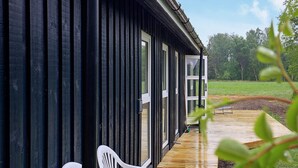 A wooden deck with two white plastic chairs, a black building with vertical siding, and a green field in the background. - 6 Person Holiday Home in Laeso (Læsø)