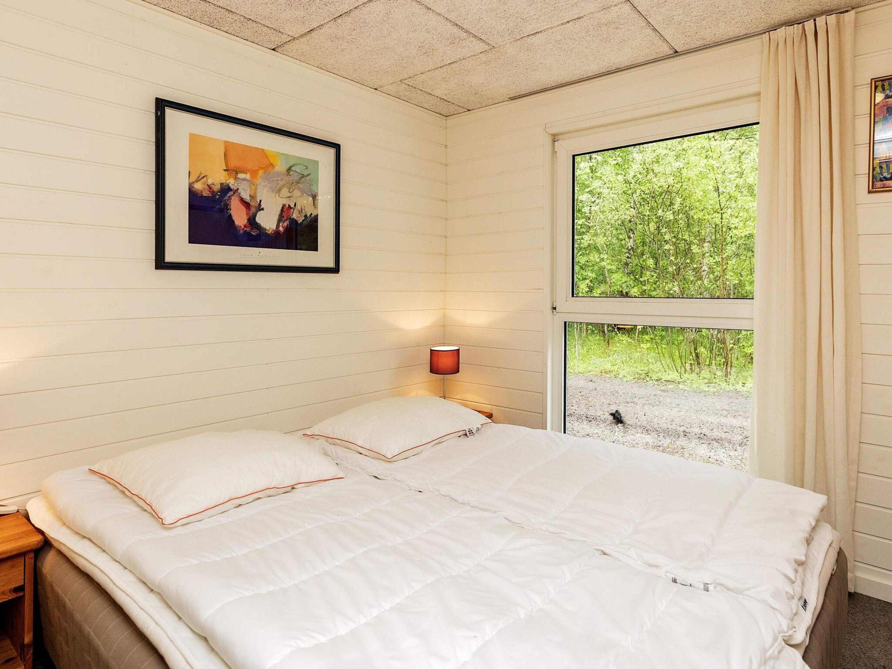 A bedroom with a bed, a nightstand, a wall-mounted lamp, a framed picture, and a window with a view of greenery.