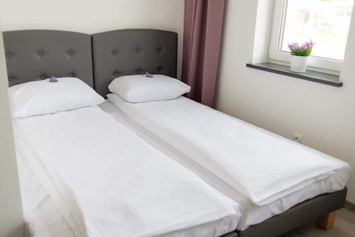 A double bed with a grey upholstered headboard, white bedding, and a window with a potted plant.
