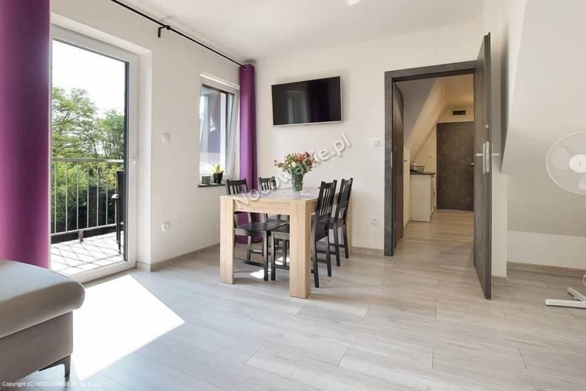 A modern dining area with a wooden table and chairs, a flat-screen TV mounted on the wall, and a view of the outdoors through large windows.