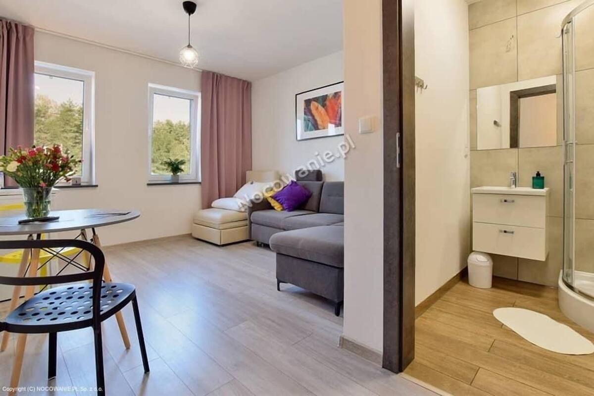 A modern living room with a sofa, a dining table with chairs, a small table with a vase of flowers, and a large window with curtains.