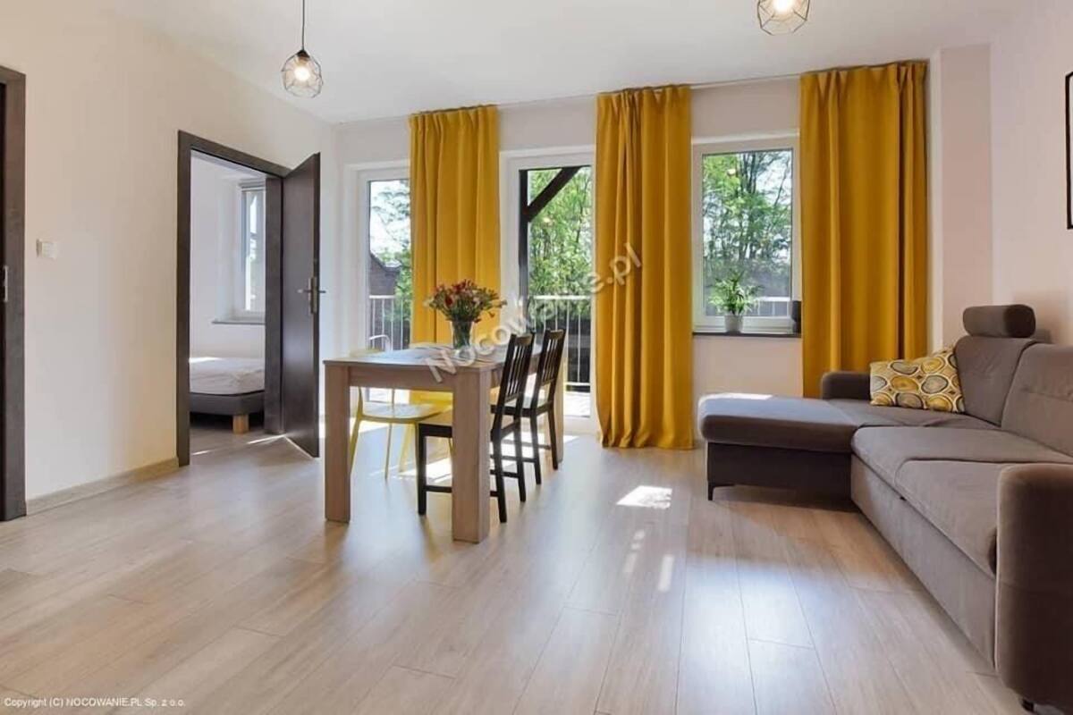 A modern living room with a sofa, dining table, and chairs. There are large windows with yellow curtains, a balcony, and a ceiling light fixture.
