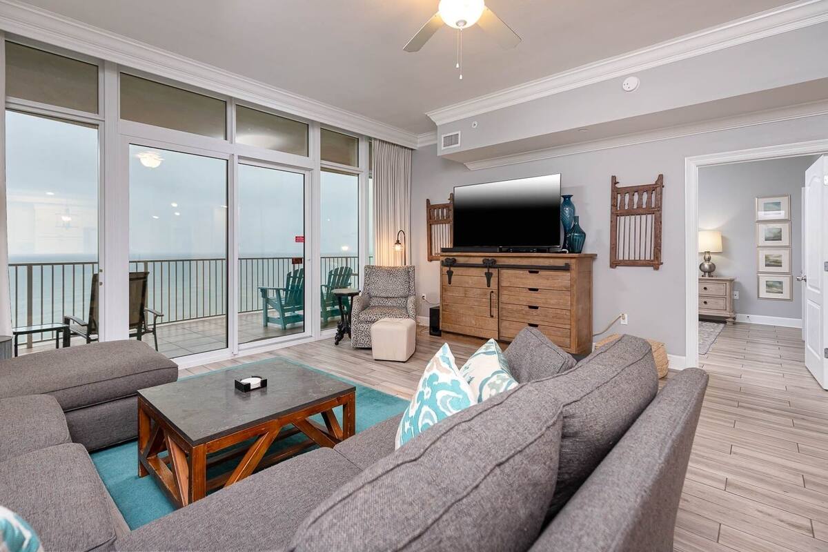 A spacious living room with a large sofa, a coffee table, and a TV on a wooden cabinet. There is a balcony with chairs and a view of the ocean.