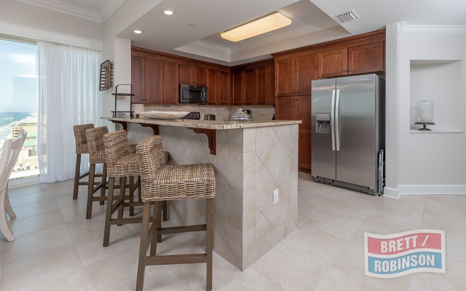A kitchen with a large island, bar stools, a refrigerator, and a view through the window.