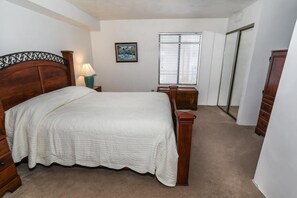 Une chambre à coucher avec un grand lit, une table de chevet, une commode, une fenêtre avec des stores et une porte.