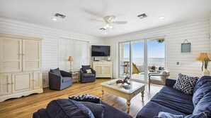 A spacious living room with a large sofa, two armchairs, a coffee table, and a television. There is a sliding glass door leading to a balcony with a view of the ocean.