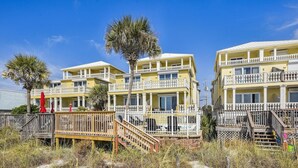 A beachfront property with multiple balconies, a wooden deck, and a sandy path leading to the entrance.