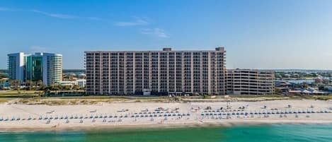 A large hotel with multiple floors and balconies, situated next to a beach with rows of umbrellas and a clear view of the ocean.