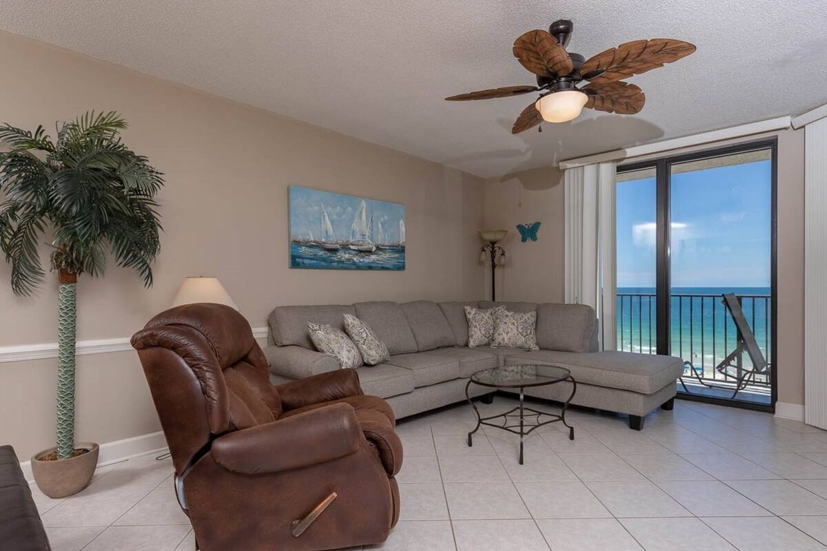Ein Wohnzimmer mit einem braunen Ledersessel, einem grauen Sofa mit gemusterten Kissen, einem Glastisch, einem Deckenventilator, einer großen Topfpflanze, einem Gemälde von Segelbooten und Meerblick durch eine Schiebetür.