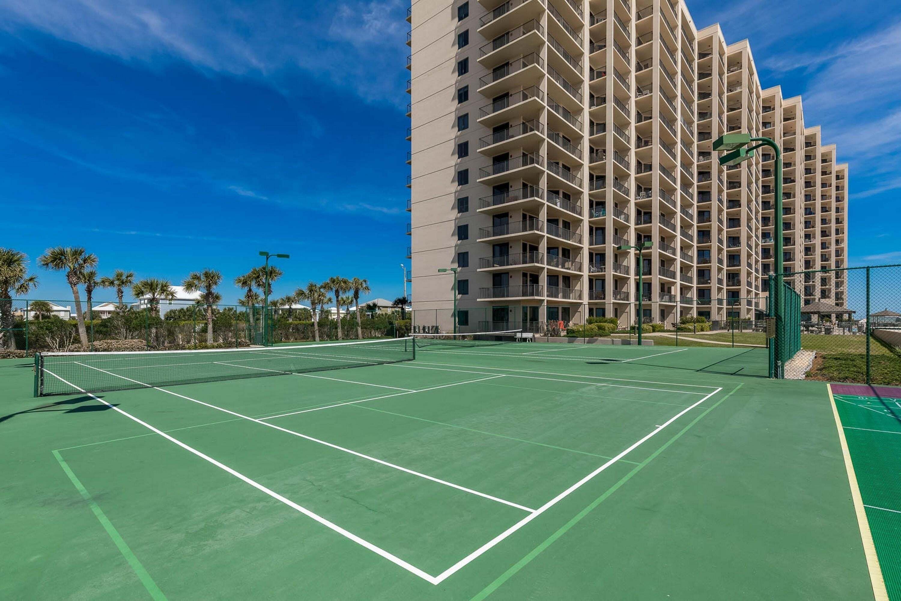 A tennis court with a net, surrounded by a fence, and a tall building in the background.
