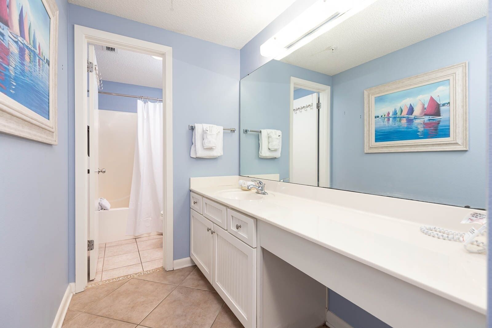 A bathroom with a white vanity, a large mirror, and a painting of sailboats on the wall.