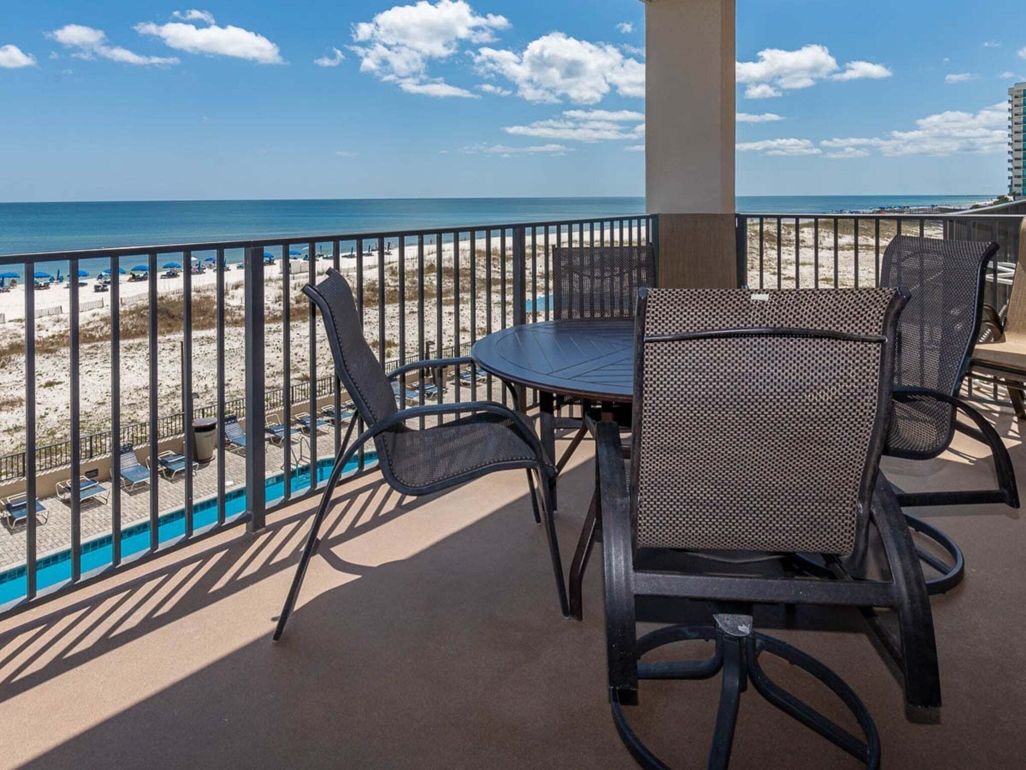 A balcony with a view of the beach, a pool, and a table with chairs.