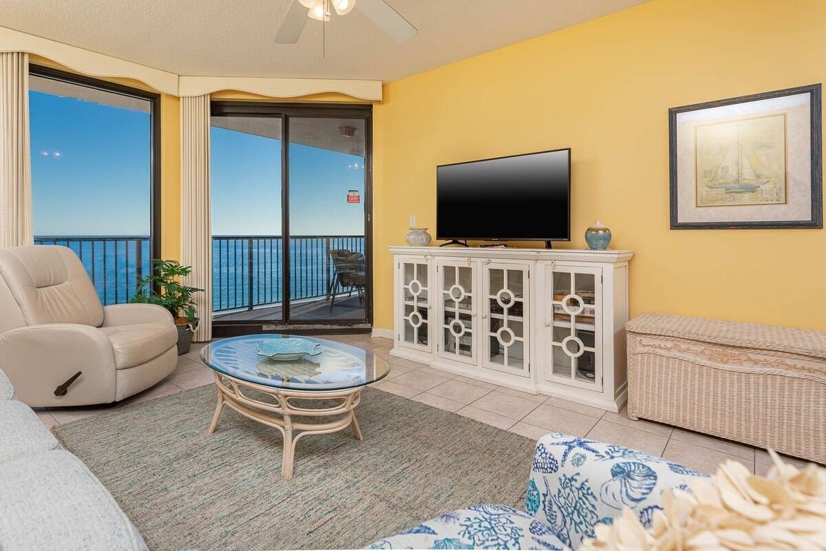 Ein Wohnzimmer mit Sofa, Sessel, Glastisch, Fernseher auf einem Schrank und einem Balkon mit Meerblick.