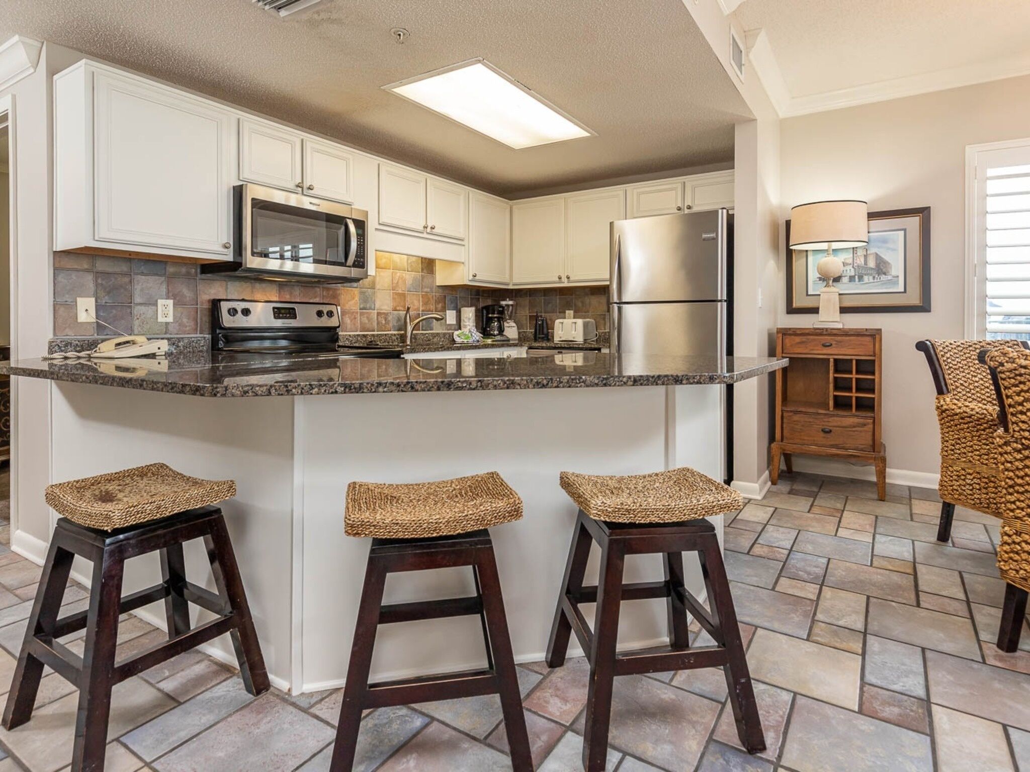 A kitchen with white cabinets, a central island with stools, a microwave, stove, refrigerator, and a coffee maker.