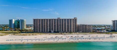 Een hotel aan het strand met meerdere verdiepingen, een zandstrand met parasols en een helderblauwe lucht.