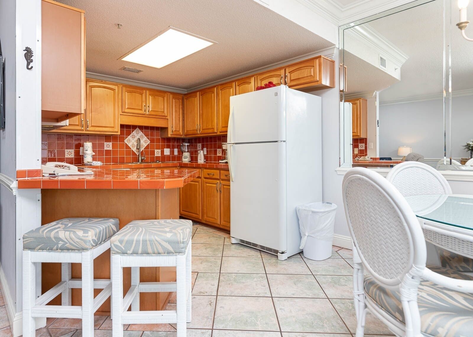 A compact kitchen with wooden cabinets, a white refrigerator, and a small dining area with white wicker chairs.