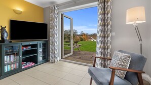 A living room with a television, a bookshelf, a chair, and a view of a garden through a sliding door. - A Family Home Near Estuary & Llansteffan Castle (Llansteffan)