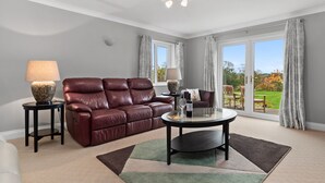 A spacious living room with a leather sofa, a glass coffee table, and a view of a garden through large windows. - A Family Home Near Estuary & Llansteffan Castle (Llansteffan)