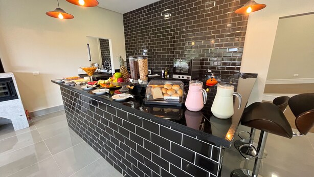 A modern kitchen with a black tiled backsplash, a central island with breakfast items, and pendant lighting. - Pousadinha Campos (Campos do Jordão)