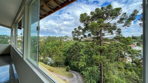 A room with a large window overlooking a lush landscape with trees and a winding road. - Pousadinha Campos (Campos do Jordão)