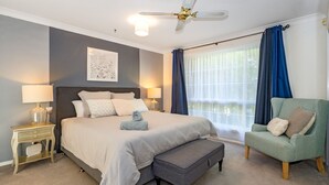 Ein Schlafzimmer mit einem Bett, Nachttischen, einem Sessel, einem Deckenventilator und einem Fenster mit Vorhängen.